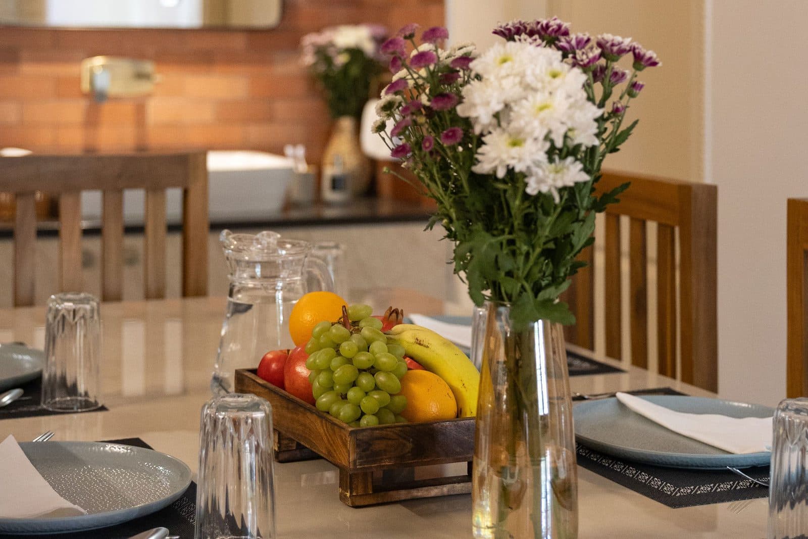 Plates, glasses, flowers and a tray of fruit on the dining table, with the wash basin behind